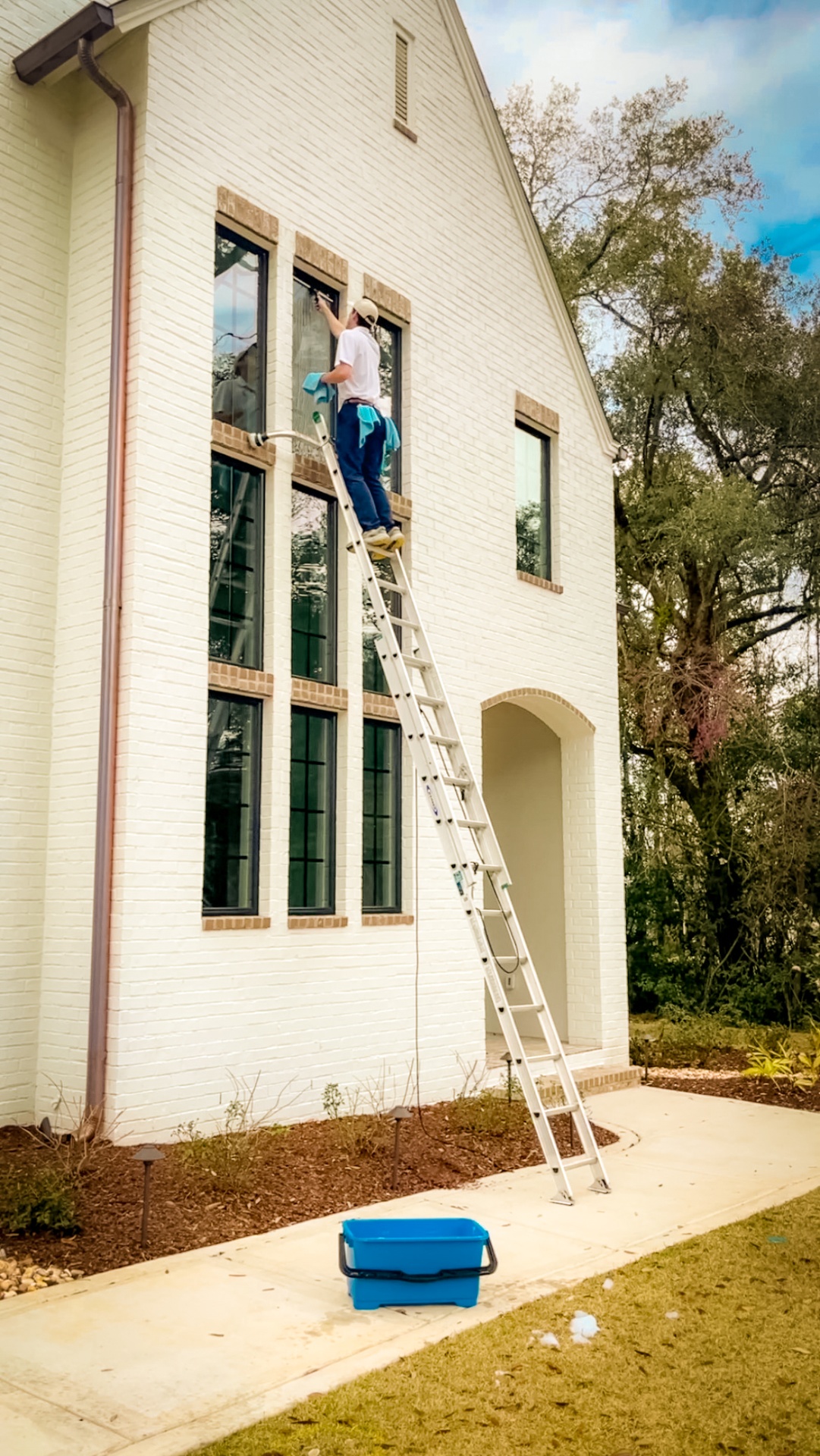 Tailored Pro Wash technician on a ladder cleaning high exterior windows of a white brick home