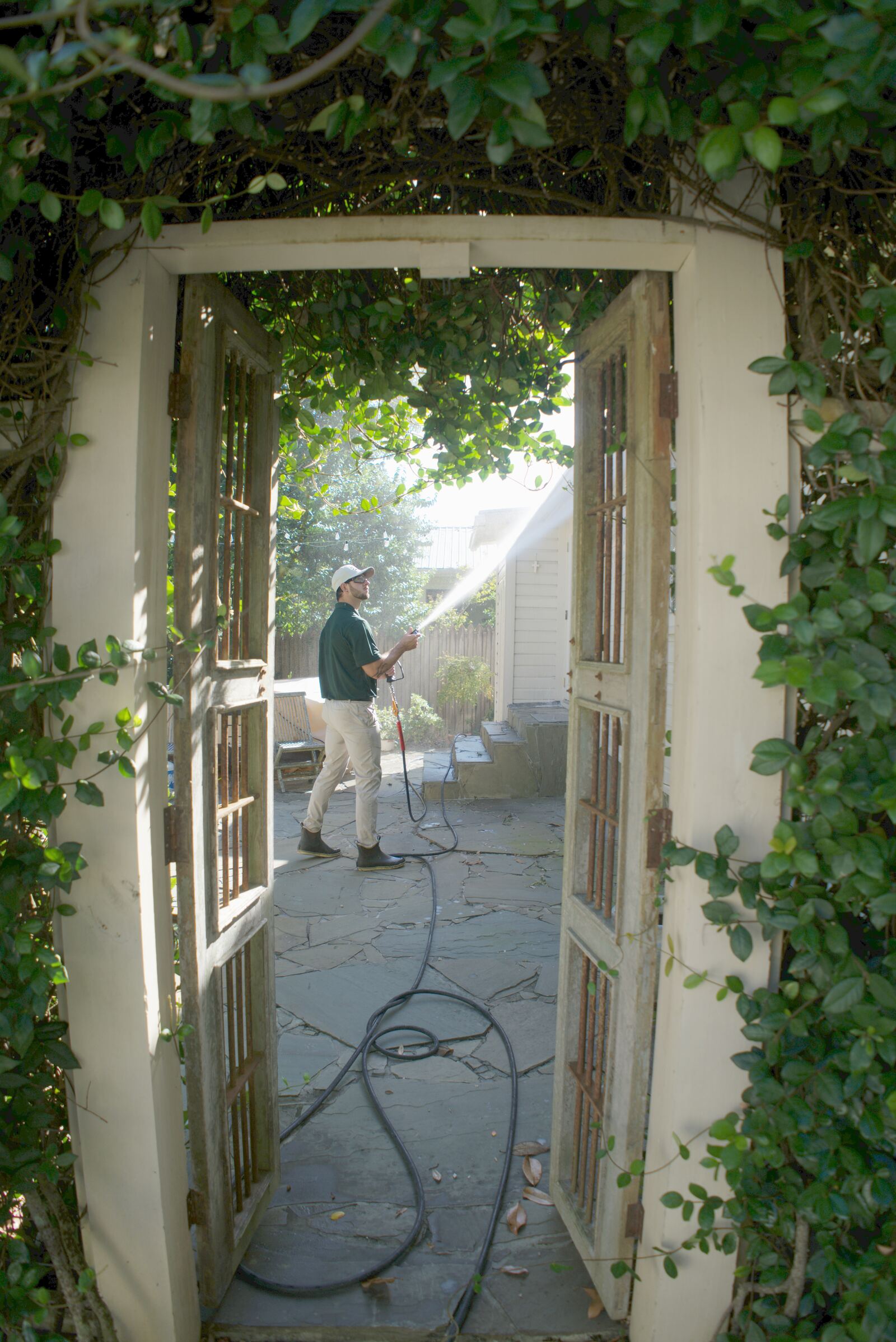 Tailored Pro Wash technician soft washing the white siding of a Fairhope, AL home, seen through a vine-covered garden gate